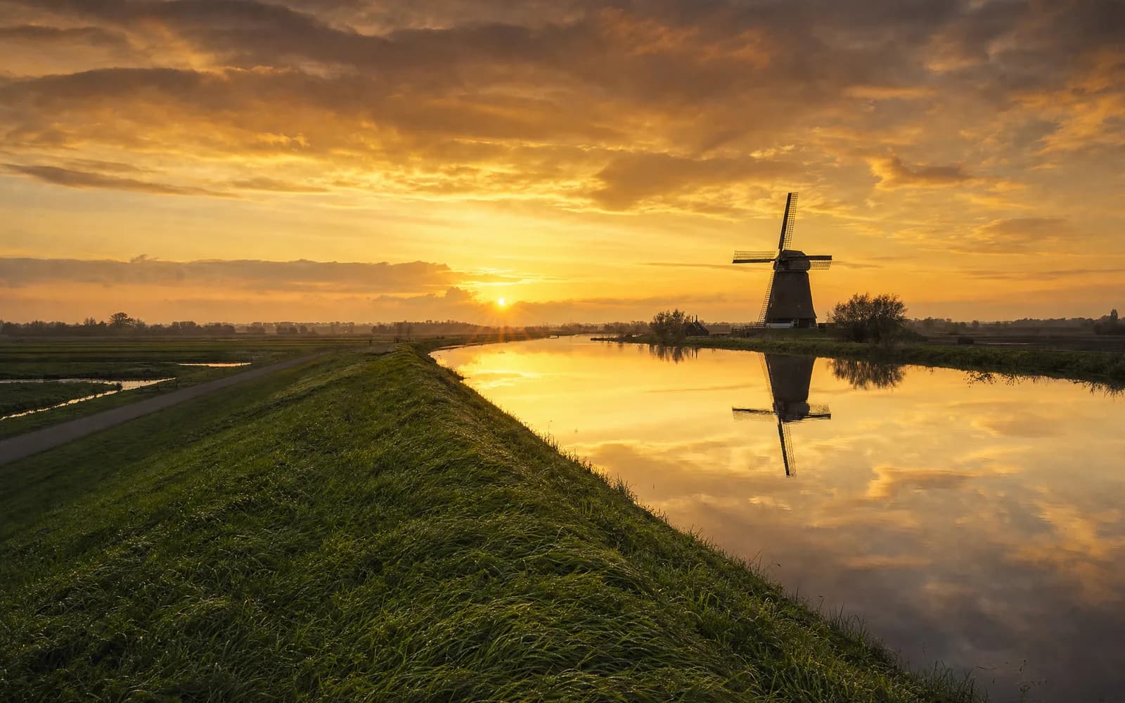 Nederlands polderlandschap met een dijk, een molen en water bij een oranje zonsondergang